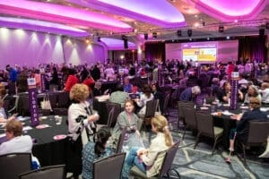 Large conference room filled with people seated at round tables, with banners for U.S. states and a screen displaying presentation slides in the background.