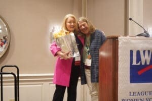 Two women stand together at a podium, one holding a gift bag and bouquet. Both are smiling. A banner with "LWV" is visible in the foreground.