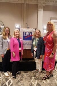 Four women stand in front of a podium with a "League of Women Voters" banner in a formal indoor setting.