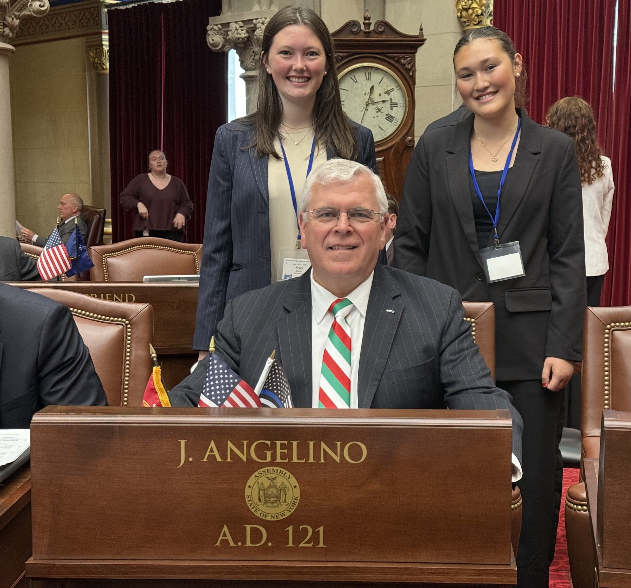 A man sits at a desk labeled "J. Angelino A.D. 121" with two young women standing behind him in a formal room with red curtains and a large clock on the wall.