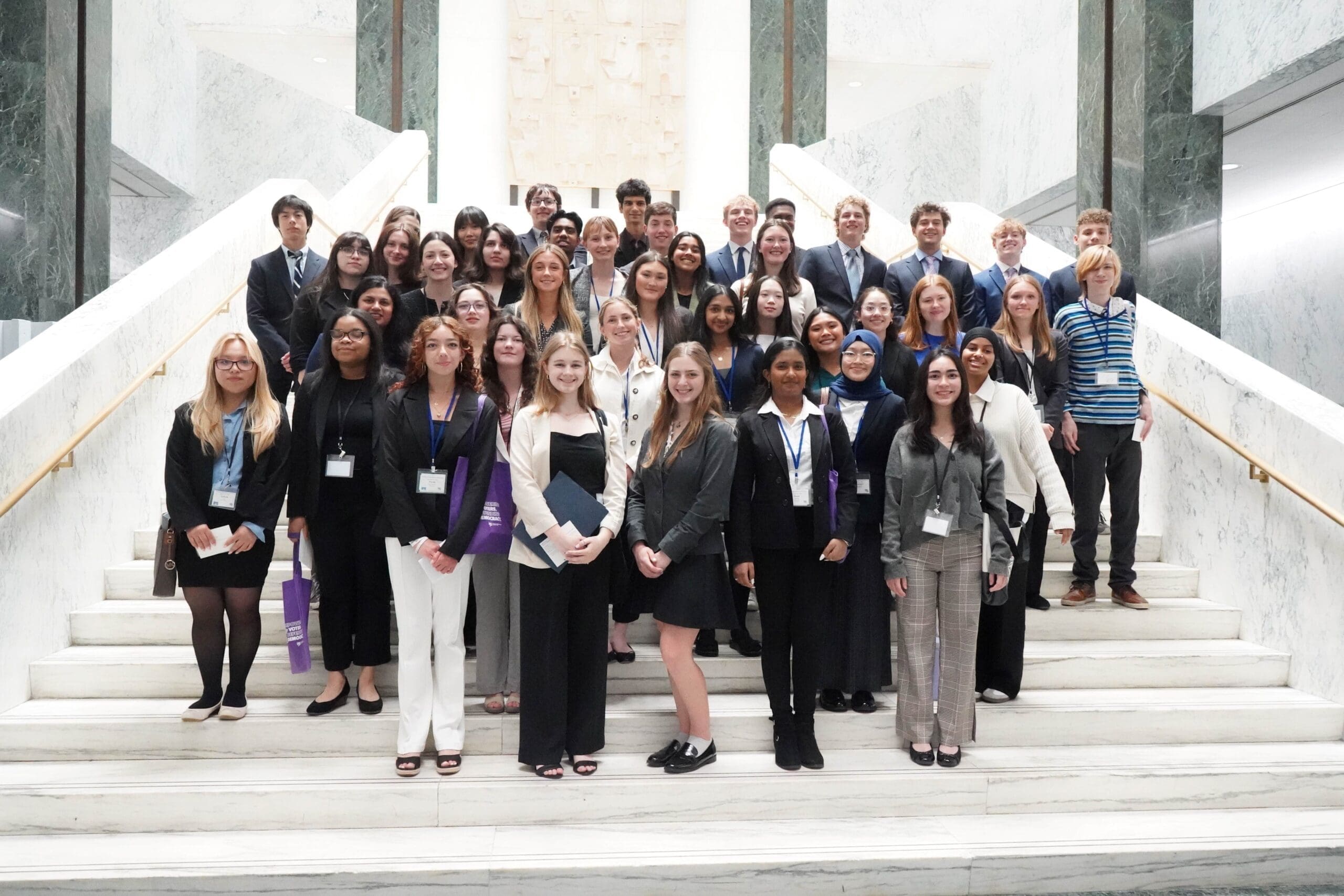 A large group of professionally dressed young adults poses on marble stairs inside a building, facing the camera and smiling.