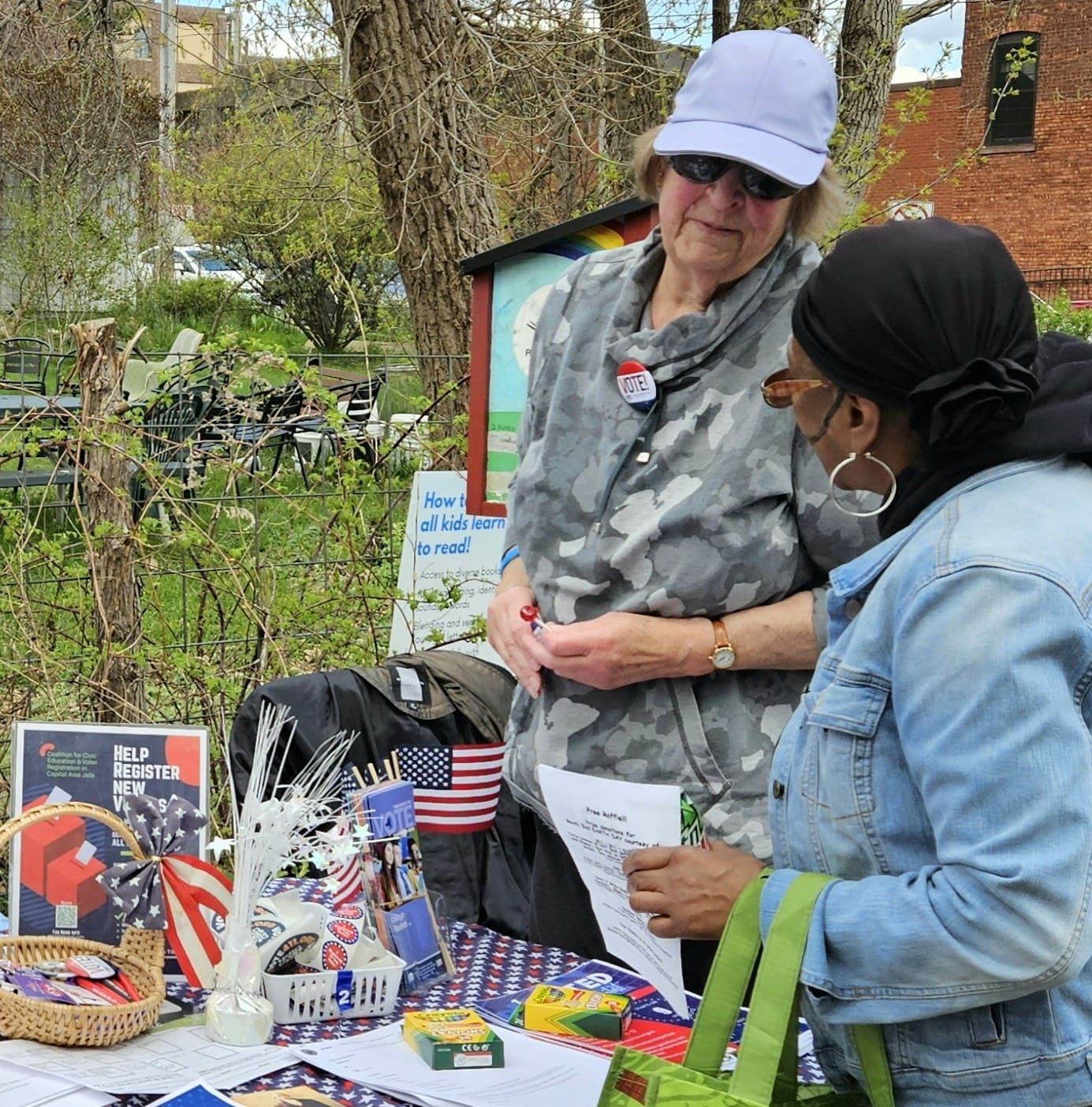 Two women stand at an outdoor table with voting information, U.S. flags, and patriotic decorations. One woman hands a paper to the other. Trees and a brick building are in the background.