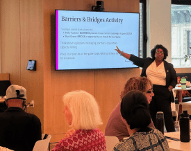 A woman stands and gestures toward a screen displaying “Barriers & Bridges Activity” as seated participants look on in a meeting room.