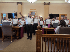 A choir of older adults, all wearing matching shirts, sings in a formal room with desks and computers, while a conductor leads them and others observe.