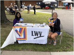 Two women sit at an outdoor table covered with an LWV banner, holding pamphlets at a community event, with other booths and people in the background.