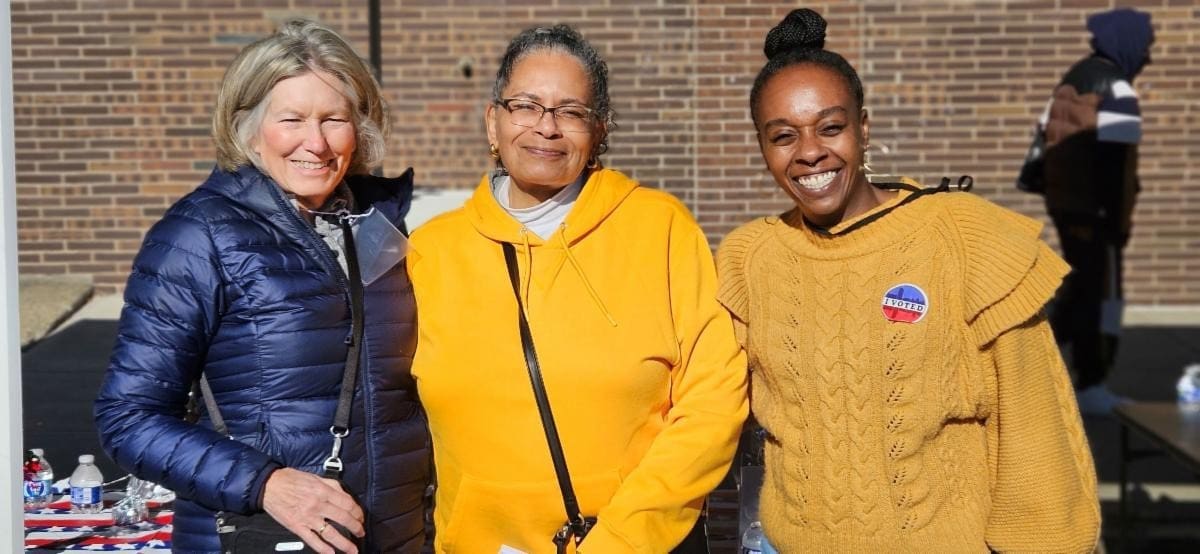 Three women stand outdoors in front of a brick wall, smiling at the camera. One wears a blue jacket, one a yellow hoodie, and one a yellow sweater with a sticker.