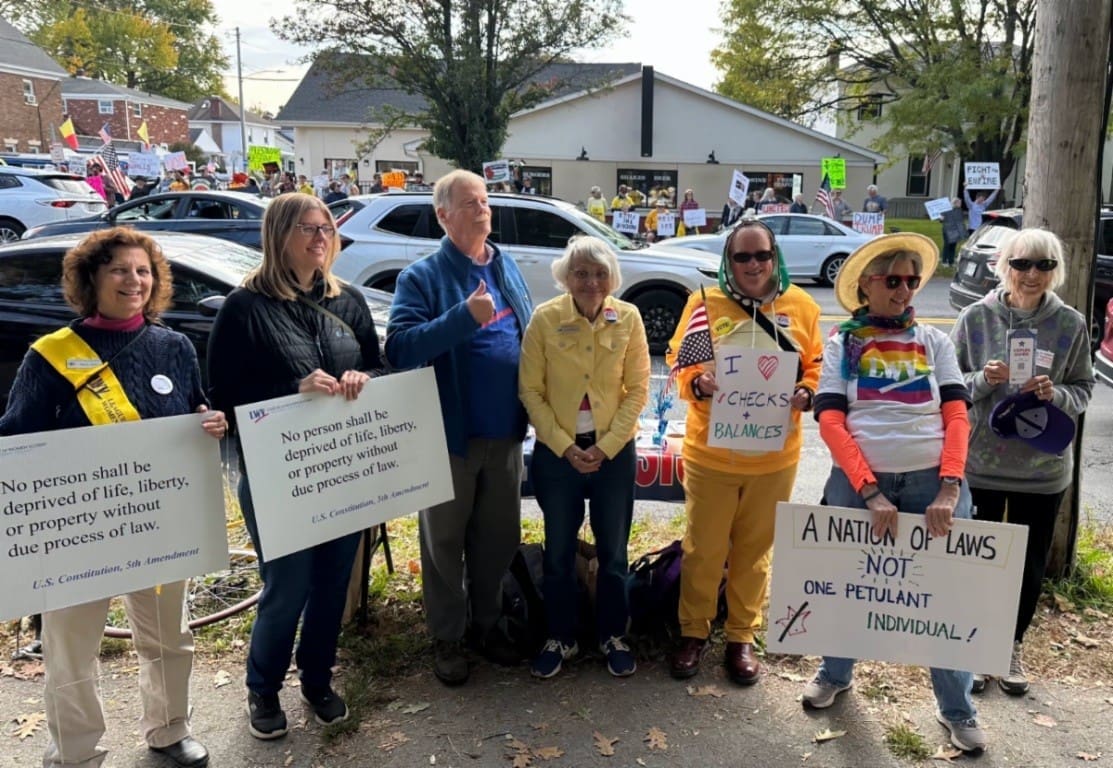 A group of seven people stand outdoors at a protest, holding signs with messages about the Constitution, checks and balances, and the rule of law. Other protesters and signs are visible in the background.