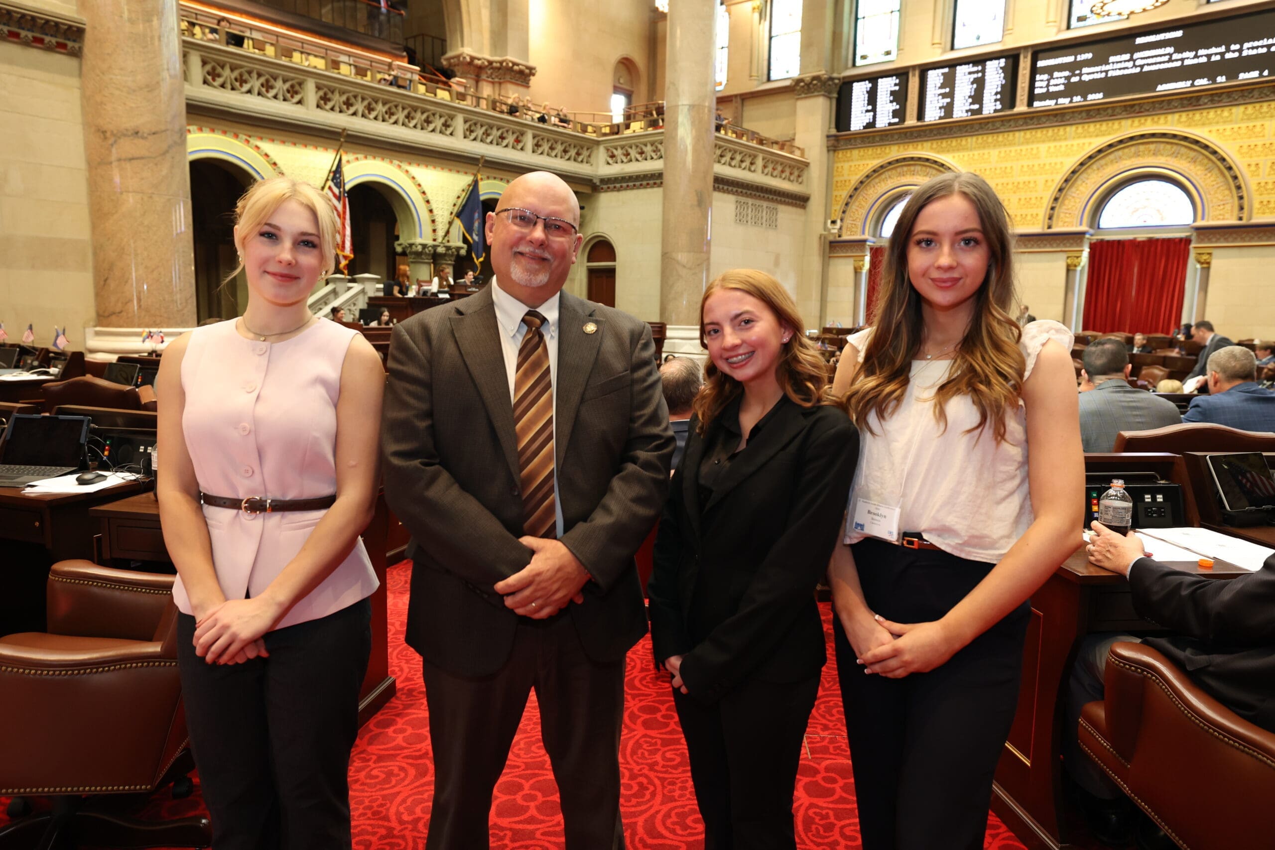 Four people stand side by side in a formal legislative chamber, three young women and one older man in business attire, with desks and a digital board in the background.