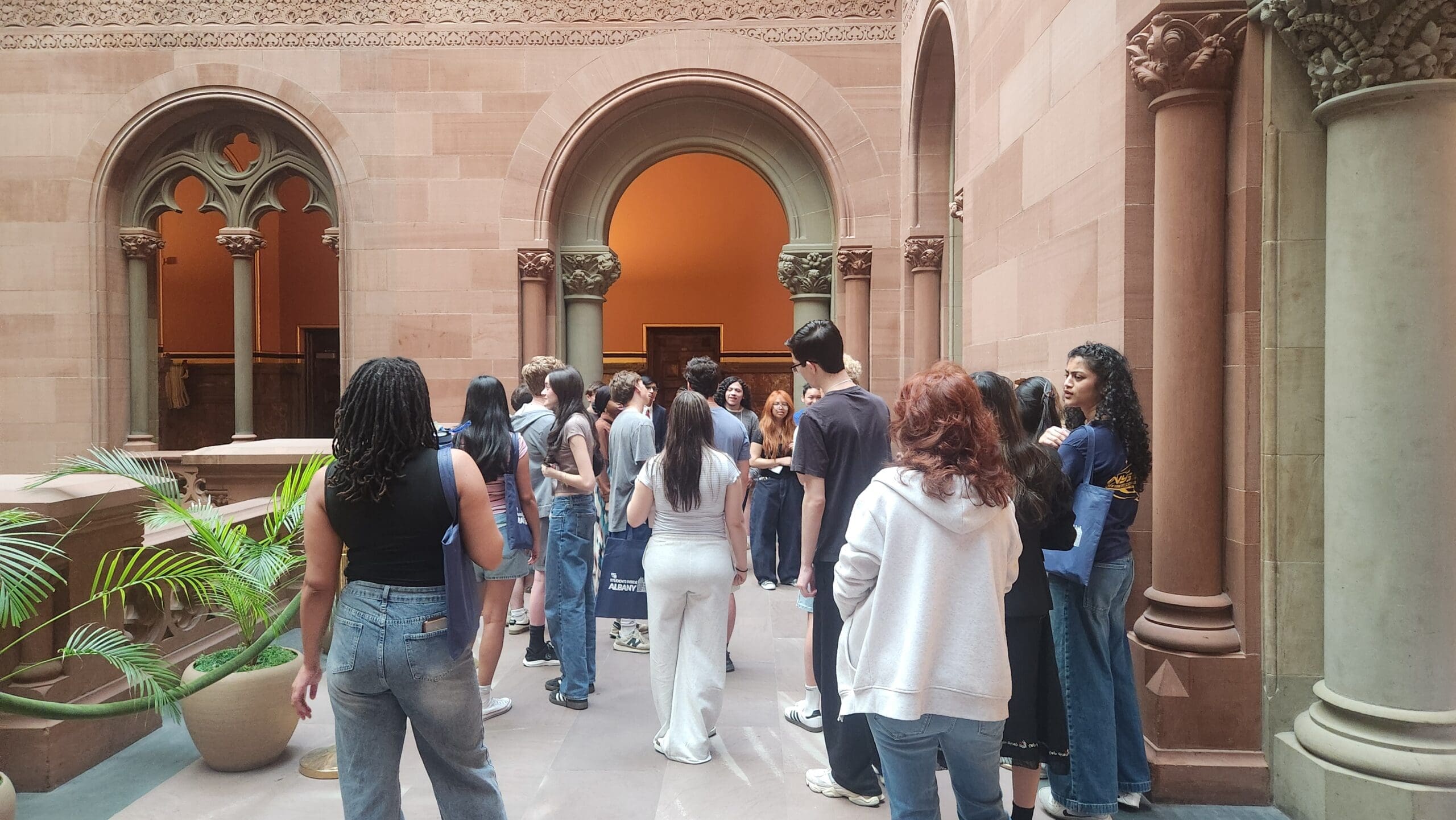 A group of people stand in a historic building with arched doorways, columns, and beige stone walls, listening to a person speak.