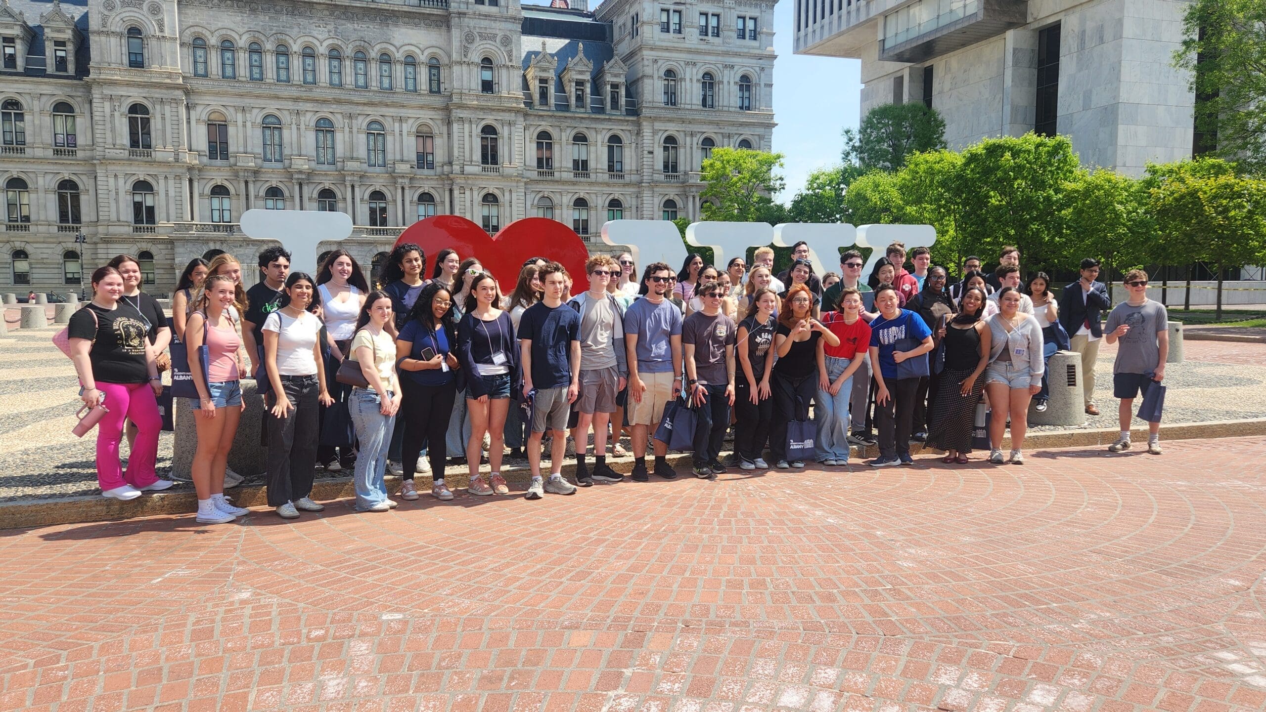 A large group of people poses in front of a sculpture with the words "I LOVE NY" in an outdoor plaza, with historic and modern buildings visible in the background.