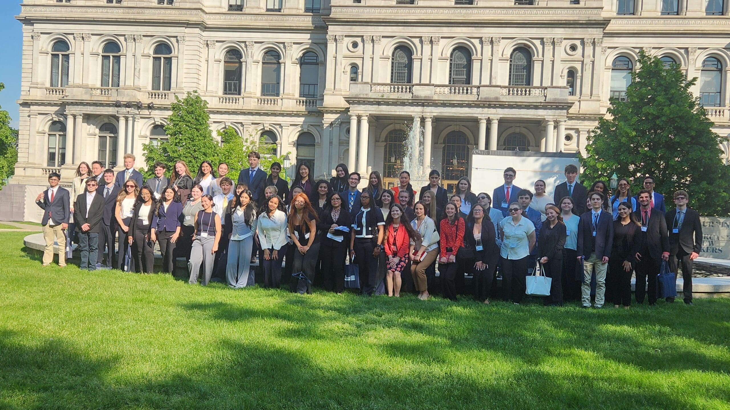 A large group of people in business attire poses for a photo outside a historic building on a sunny day.