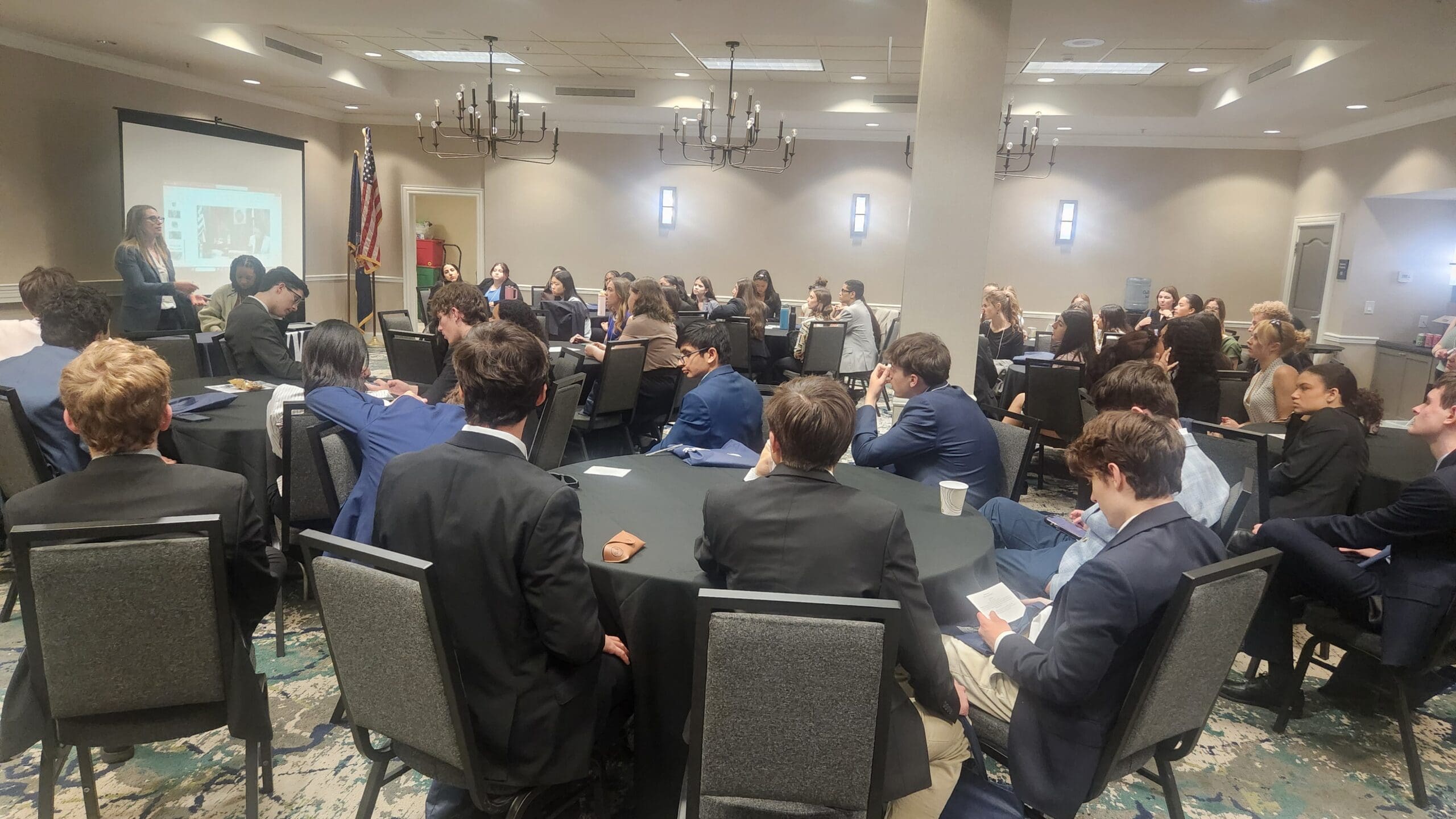 A group of people in business attire sit at round tables in a conference room, listening to a presenter at the front near a projection screen.