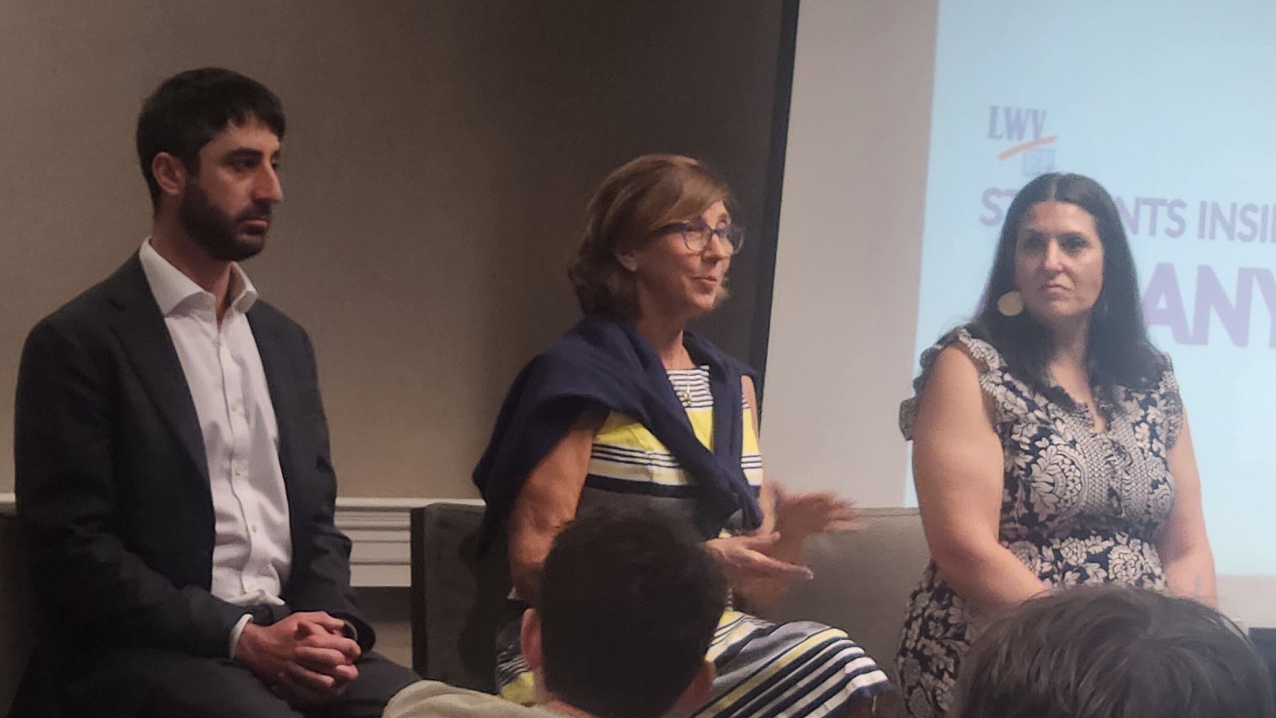 Three adults sit on a panel in front of an audience; a projection screen with partial text is visible in the background.