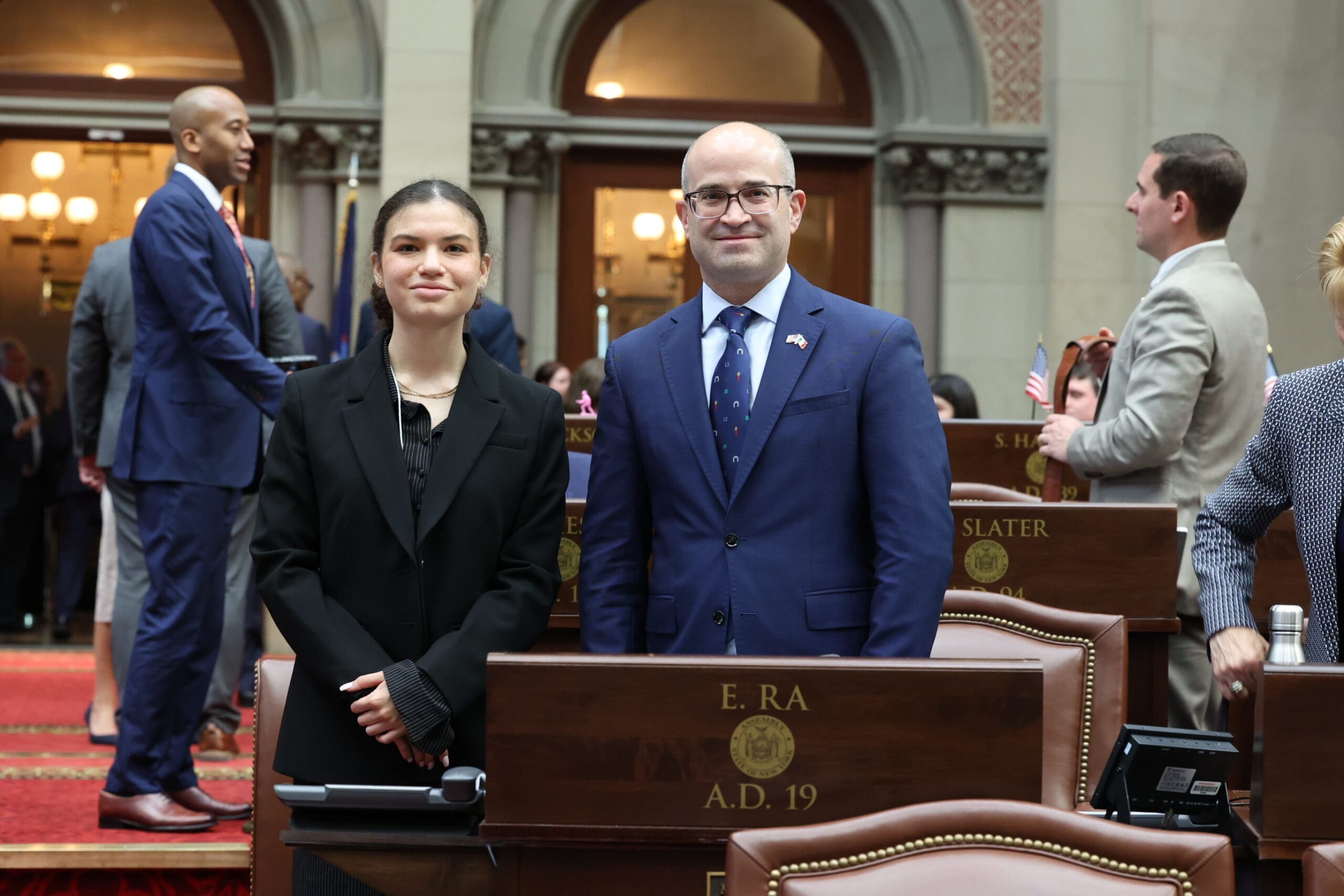 Two people in formal attire stand in a legislative chamber with desks and other people in the background.