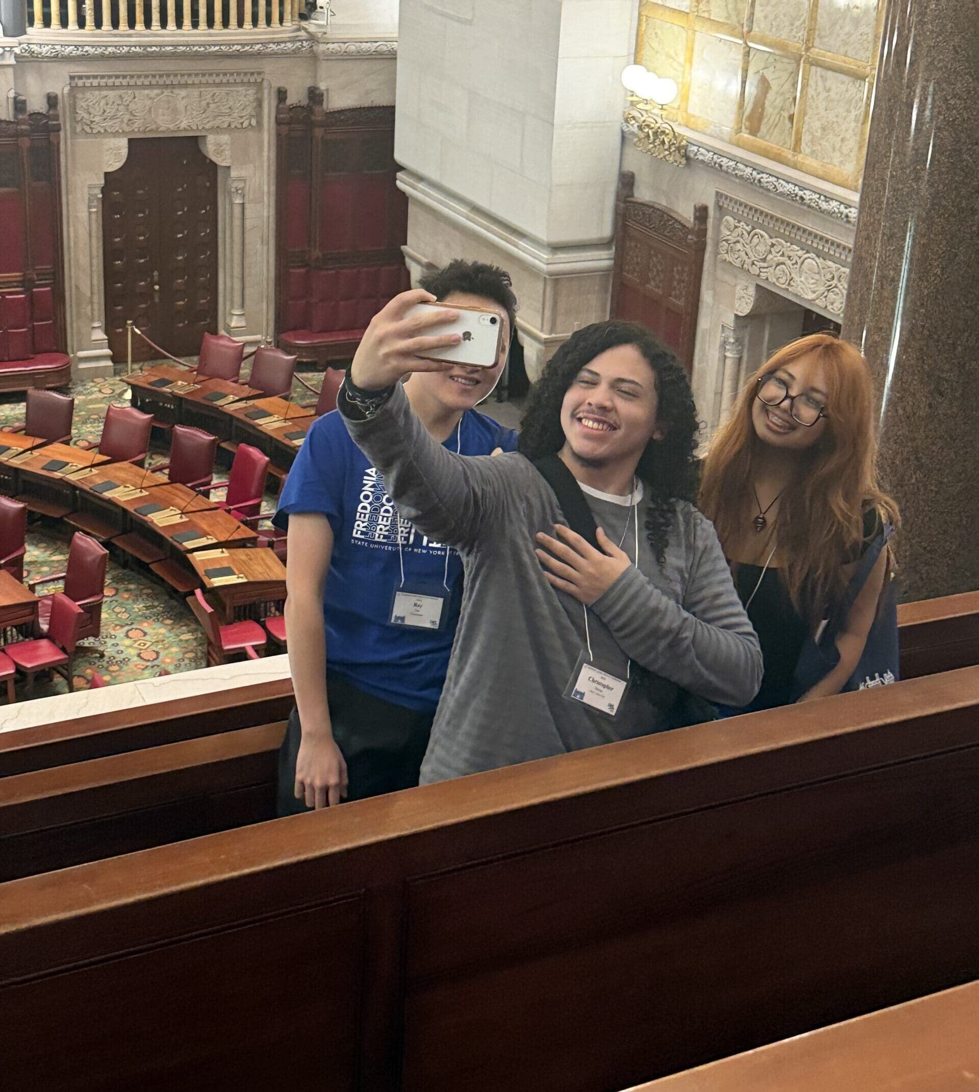 Three young people take a selfie in front of a senate chamber with red seats and ornate architecture.