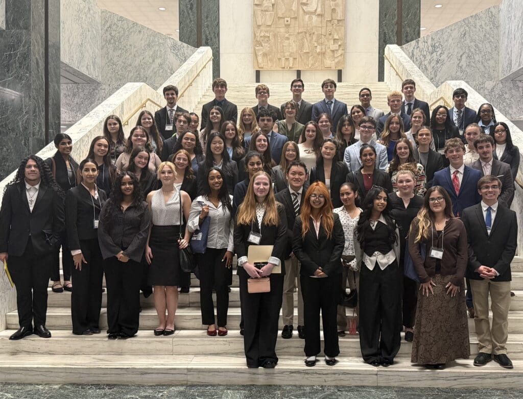 A large group of professionally dressed students stands on marble stairs in a formal building, posing for a group photo.