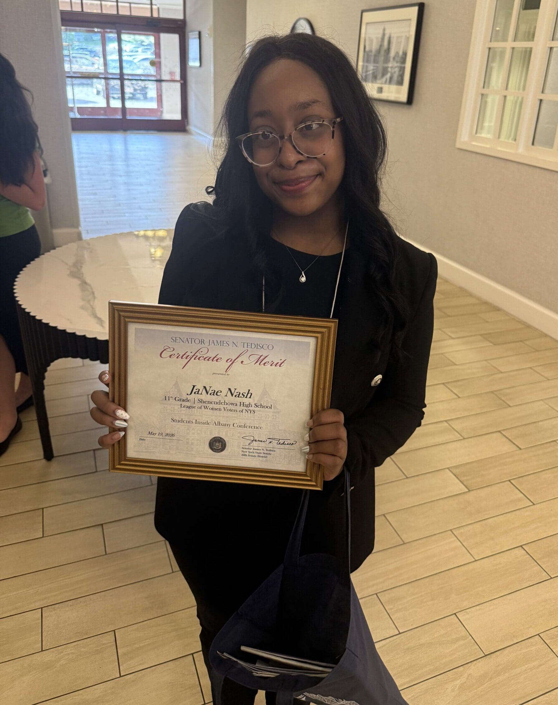 A woman wearing glasses and a black outfit holds a framed certificate of merit while standing indoors on a light-colored tiled floor.