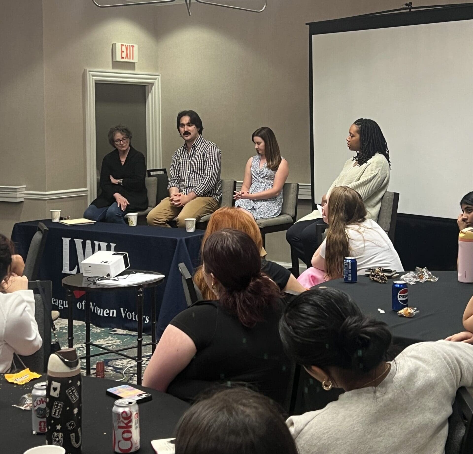 Four people sit at the front of a conference room, speaking to an audience. A table with drinks and snacks is in the foreground. A "League of Women Voters" banner is visible.